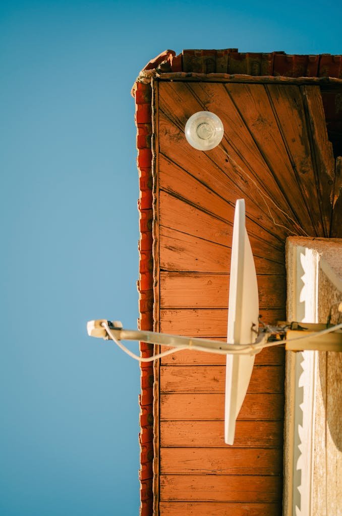 Gros plan sur un toit en bois rustique où se dresse une antenne parabolique, sur fond de ciel bleu azur. Tons et textures chauds.