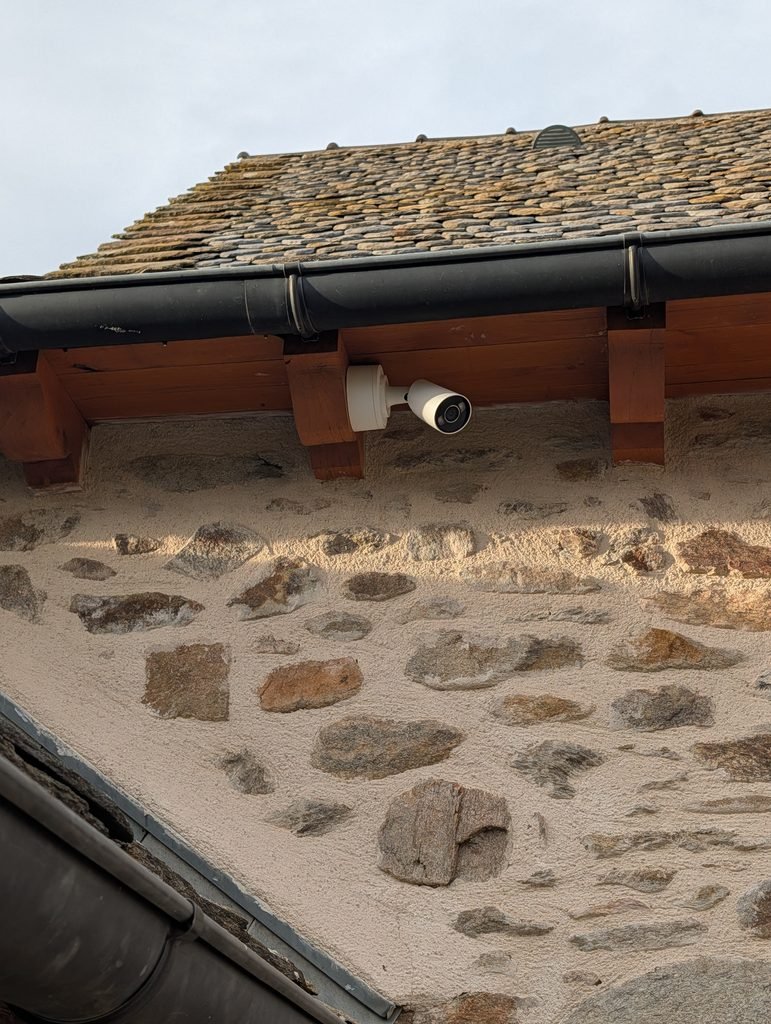 Caméra de vidéosurveillance extérieure Ajax avec éclairage LED, installée sous une avancée de toiture sur une façade en pierre par JOUVE SCF.