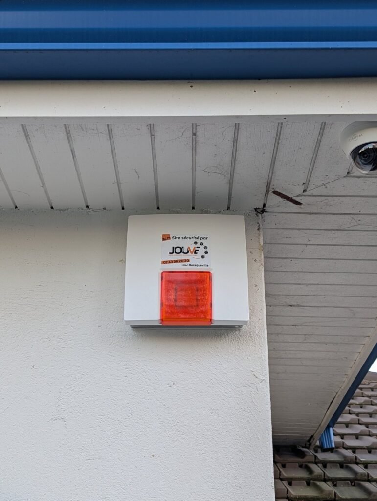 Sirène d’alarme extérieure Daitem avec flash lumineux orange, installée en façade sous une avancée de toiture par JOUVE SCF pour sécuriser un bâtiment.